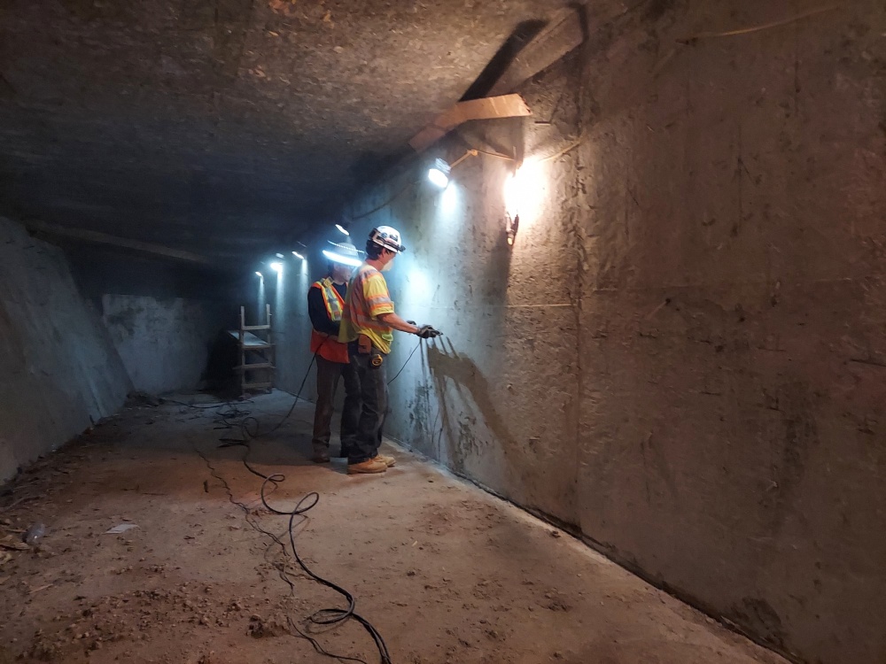 Two people wearing PPE in a box girder using a Impact Echo/Pulse Velocity device to evaluate the structures reinforcements within the concrete.